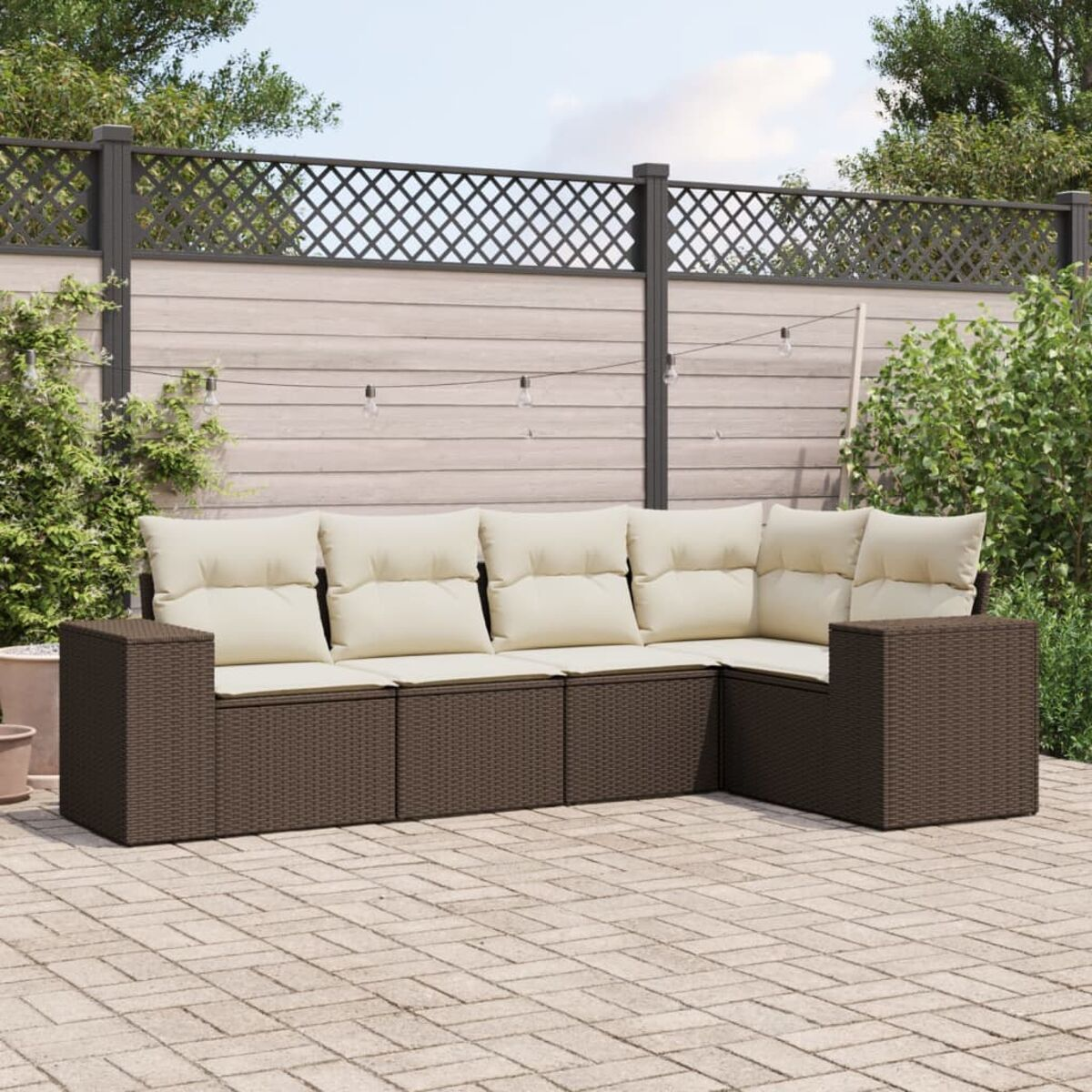 Divano angolare da esterno a forma di L in vimini marrone con cuscini beige, su un patio in mattoni, con recinzione e vegetazione.