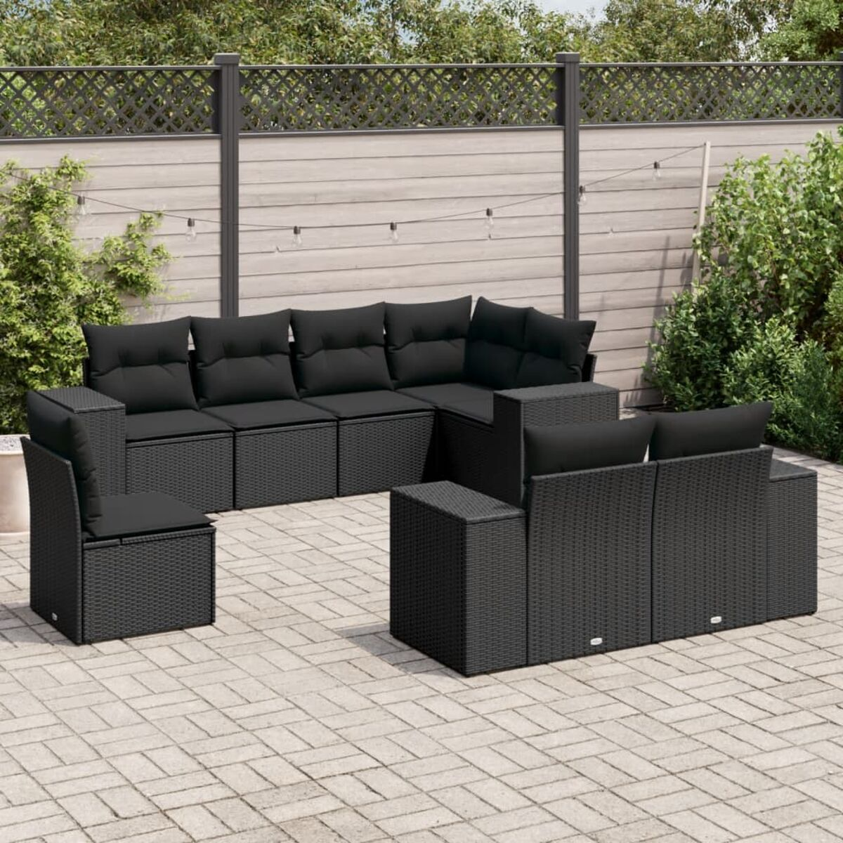 Schwarze Rattan-Terrassenmöbel. Schwarze Kissen, Holztisch. Grüne Pflanzen.