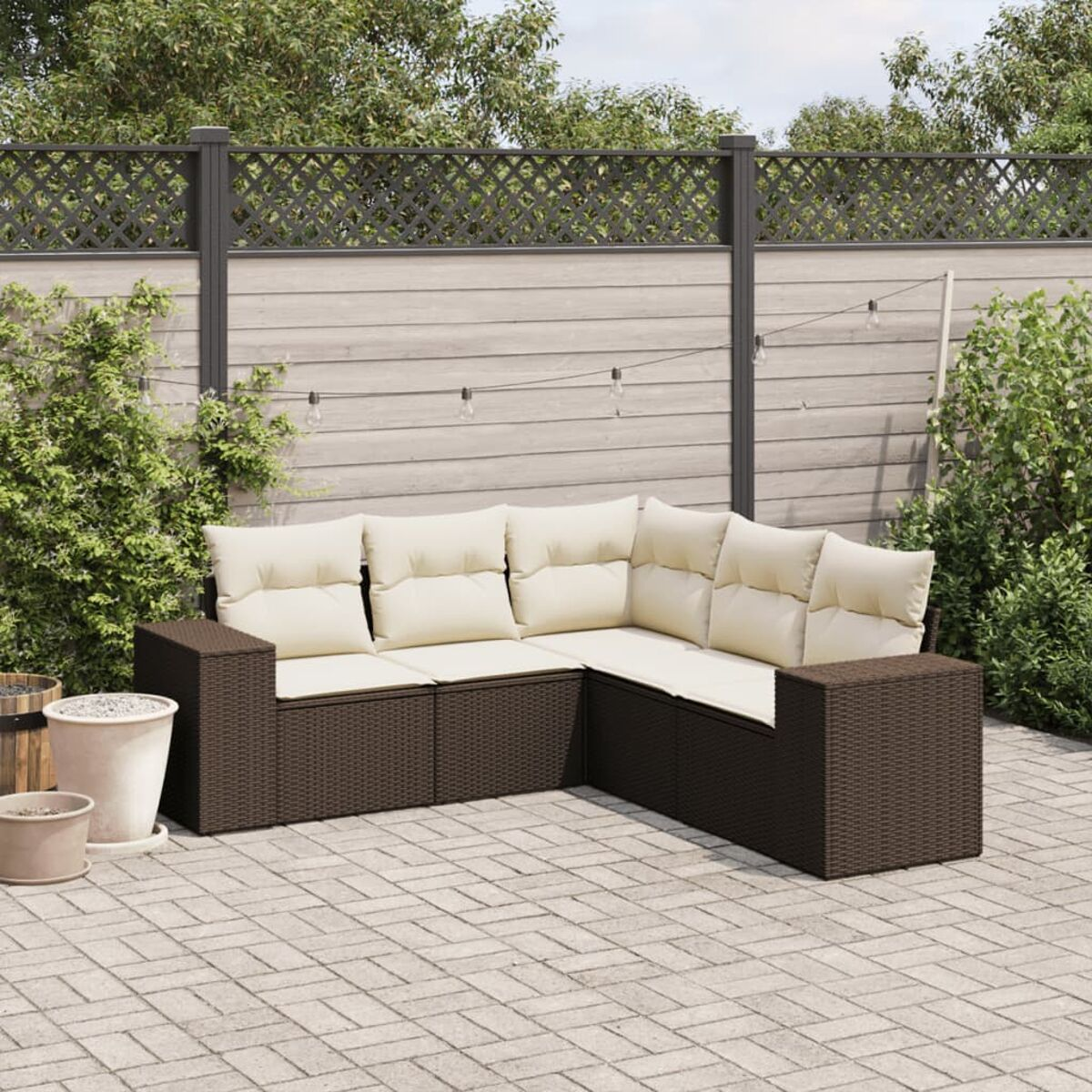 Divano componibile da esterno in vimini marrone con cuscini beige, su un patio in mattoni, con verde e recinzione.