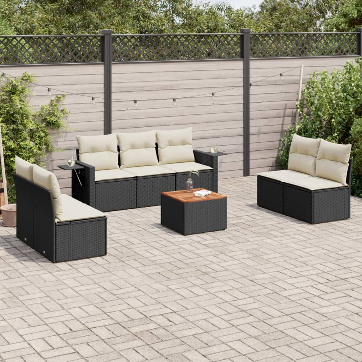 Schwarzes Korbmöbel-Set für die Terrasse mit cremefarbenen Kissen und einem Holztisch auf einer gepflasterten Terrasse. Grün und Zaun im Hintergrund.