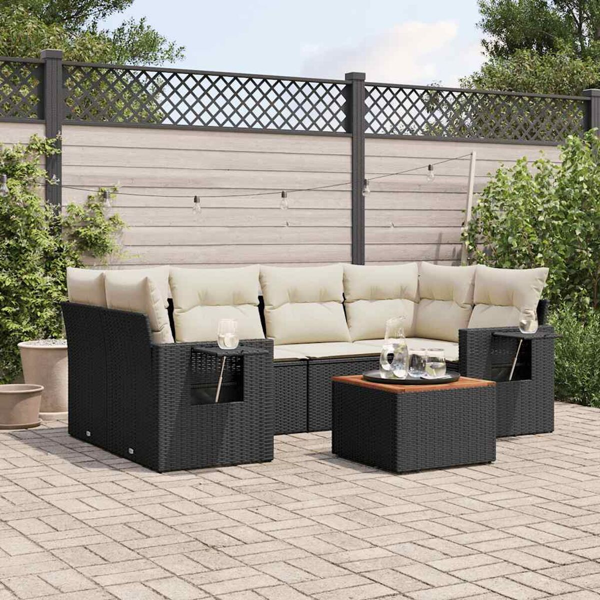 Schwarzes Rattan-Sofa-Set mit cremefarbenen Kissen, Holztisch und Lichterketten an einem Holzzaun.