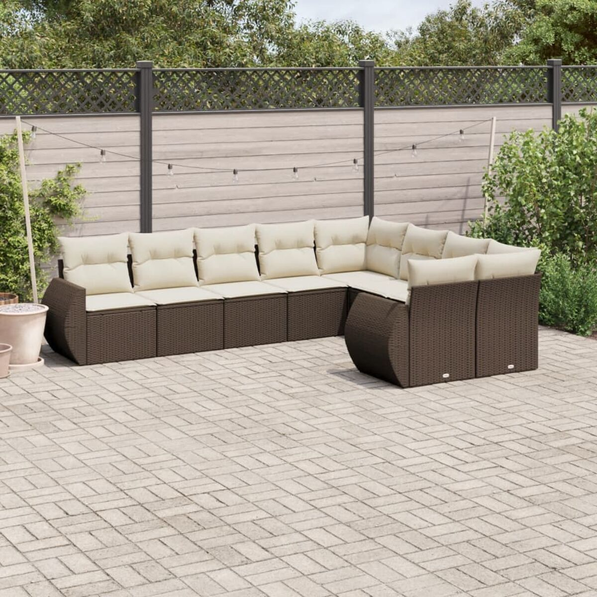 Divano componibile da esterno in vimini marrone con cuscini beige su un patio pavimentato. Piante verdi e una recinzione sullo sfondo.
