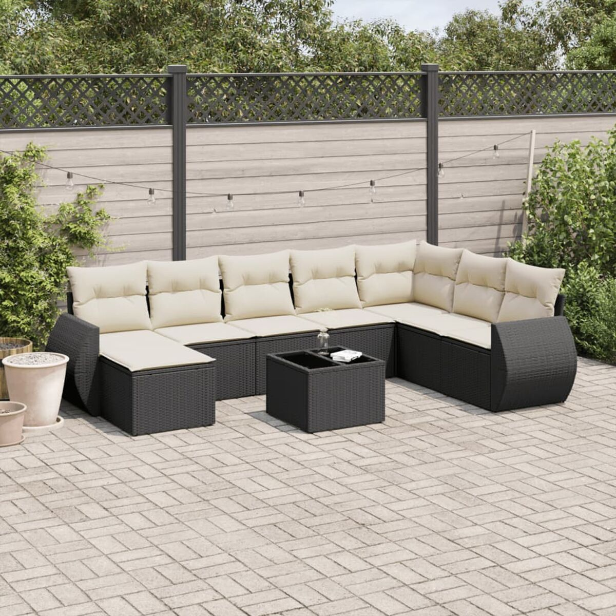 Schwarzes Rattan-Ecksofa mit beigen Kissen auf einer Terrasse mit Zaun und Pflanzen.