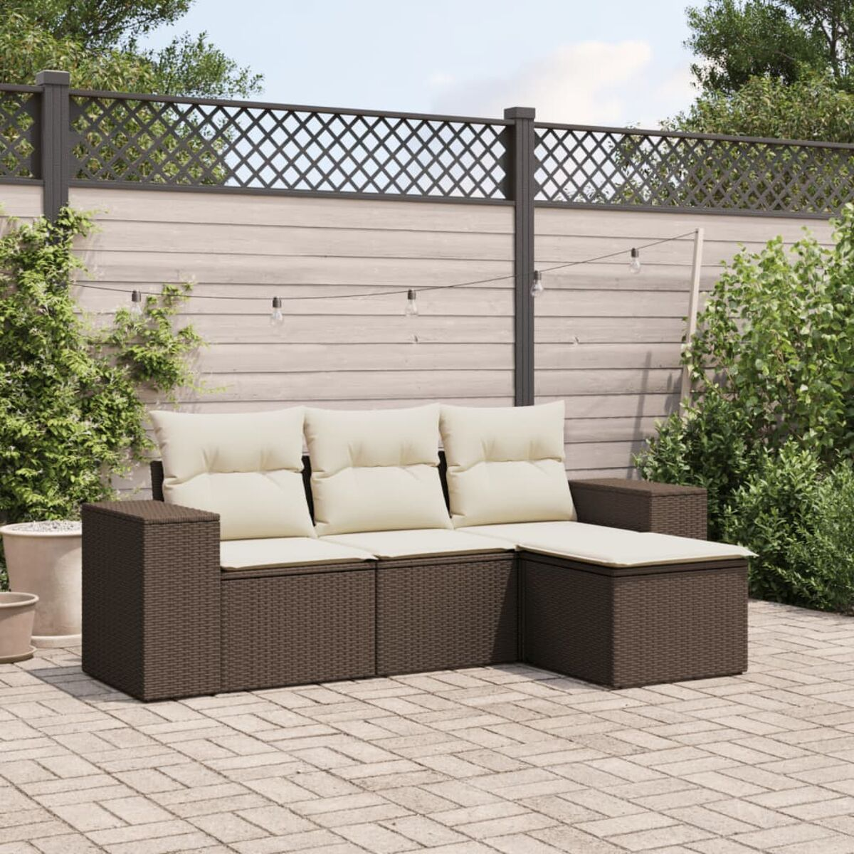 Divano angolare da esterno con struttura in vimini marrone e cuscini beige. Patio in mattoni. Recinzione in legno sullo sfondo.