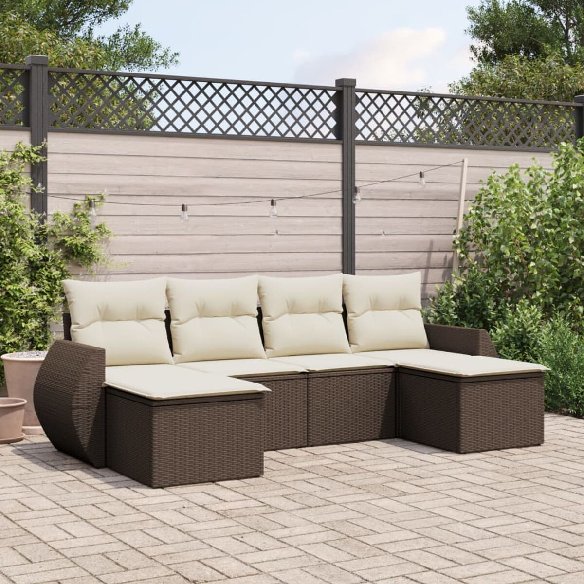 Divano componibile da esterno in vimini marrone con cuscini color crema su un patio in mattoni, una recinzione e verde sullo sfondo.
