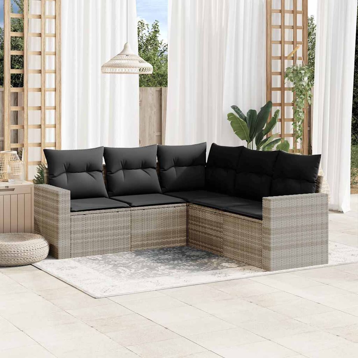 L-förmiges Rattansofa mit schwarzen Kissen auf einem Teppich, im Freien. Weiße Vorhänge und Pflanzen im Hintergrund.