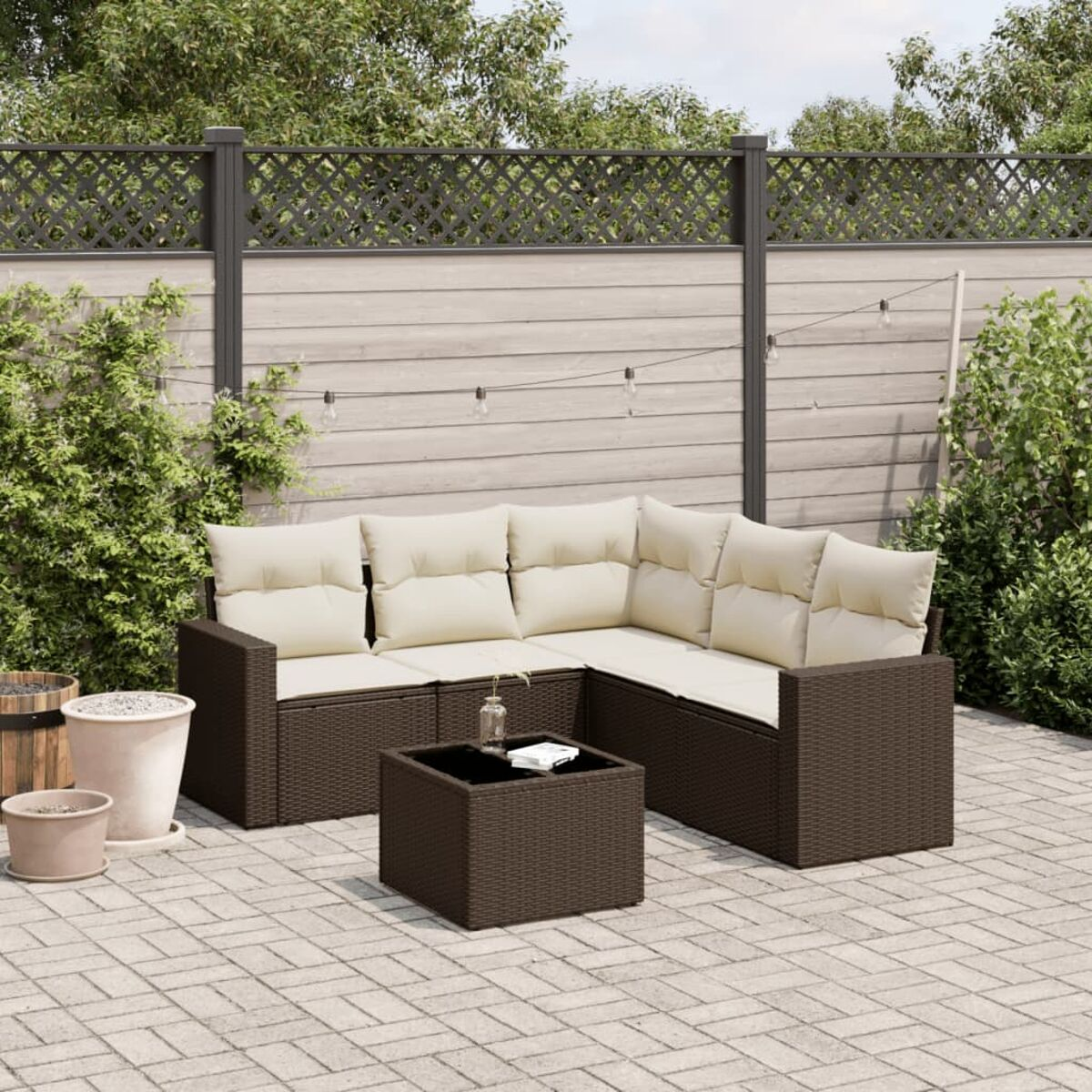 Divano in rattan marrone con cuscini color crema e tavolo nero su patio in mattoni con verde.