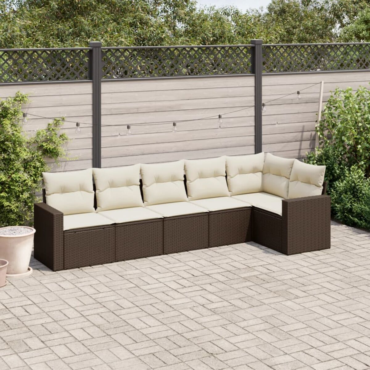 Dunkelbraune geflochtene Outdoor-Eckcouch mit cremefarbenen Kissen. Auf einer Steinterrasse. Zaun und Sträucher.