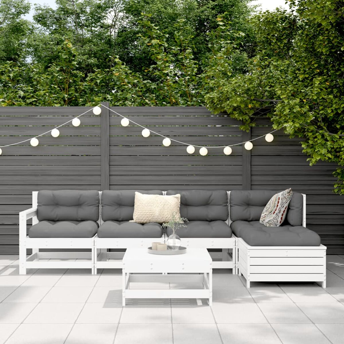 Weißes Holz-Ecksofa mit grauen Kissen und weißem Tisch. Hintergrund: Zaun und Lichter.