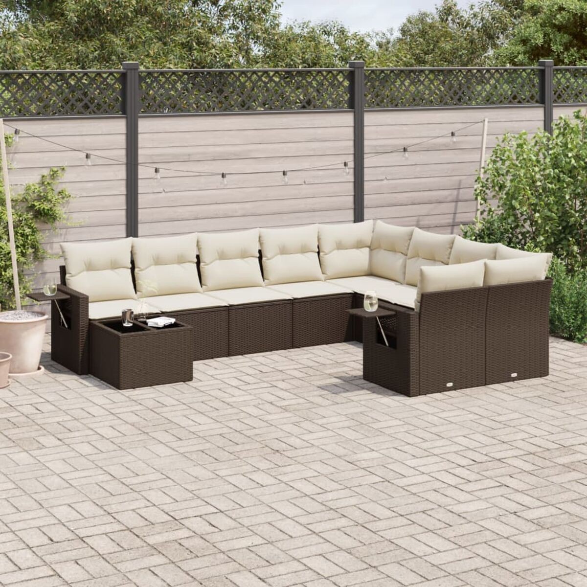 Divano angolare da esterno in vimini marrone con cuscini color crema, tavolini, su un patio. Recinzione e verde sullo sfondo.