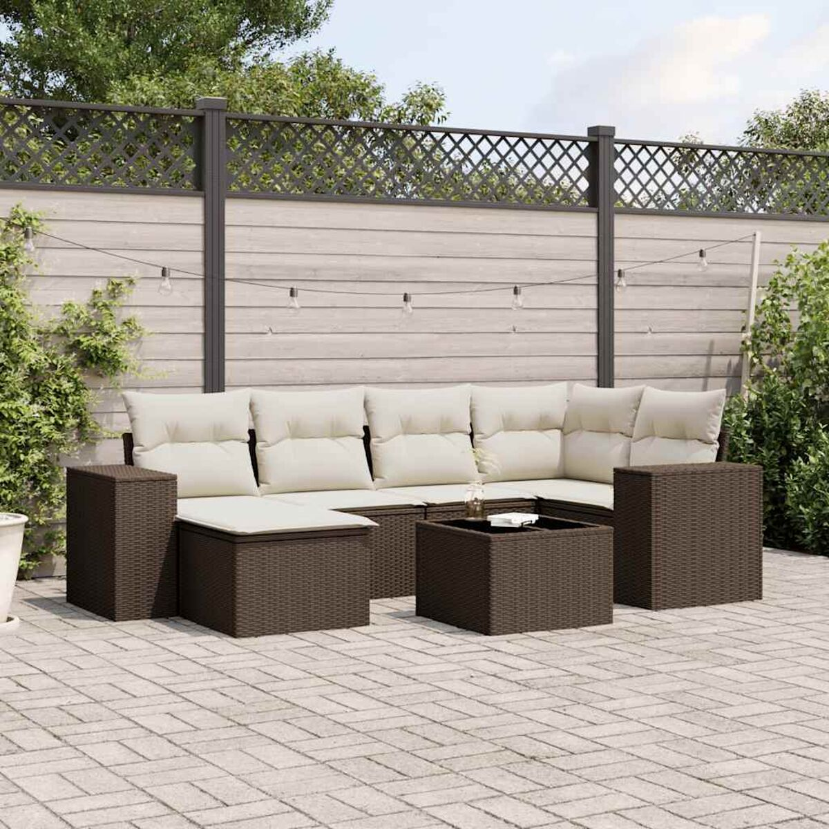Divano angolare da esterno in vimini marrone con cuscini beige e due tavolini, su un patio in mattoni con recinzione in legno.