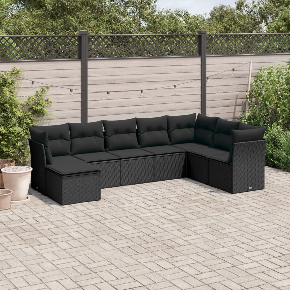Schwarzes Outdoor-Ecksofa mit schwarzen Kissen auf einer Backstein-Terrasse. Zaun und Grün im Hintergrund.