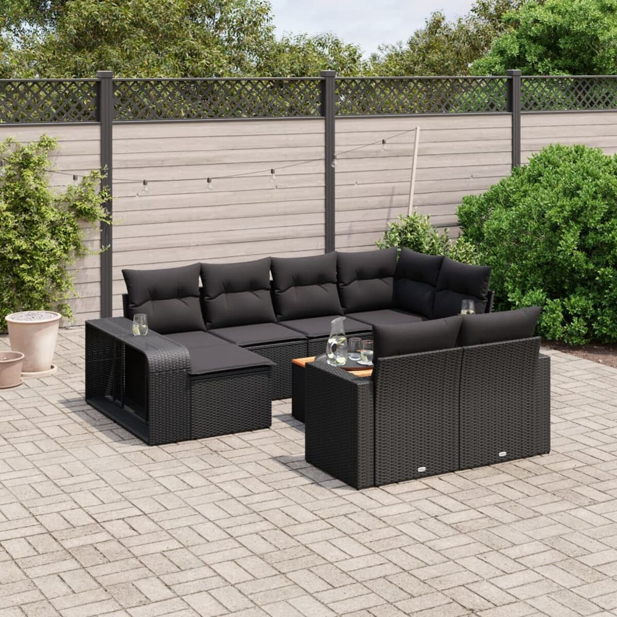 Schwarzes Outdoor-Ecksofa aus Korbgeflecht mit schwarzen Kissen auf einer Backstein-Terrasse, mit Glas und Pflanzen.