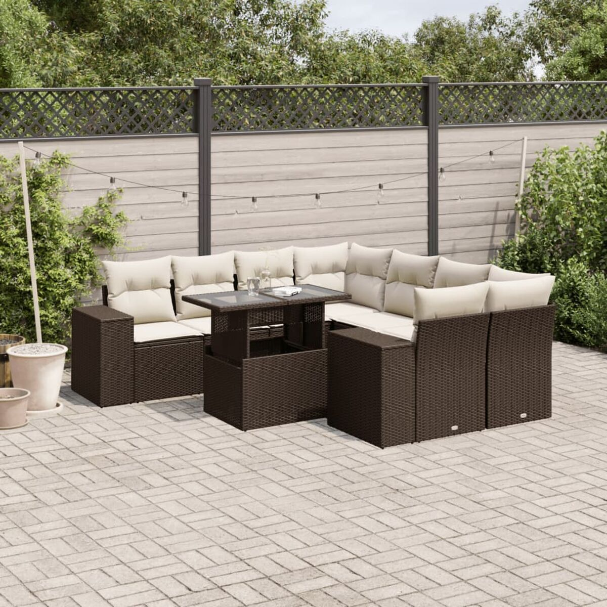Divano angolare in vimini marrone con cuscini beige e tavolino su un patio in mattoni. Piante verdi sullo sfondo.