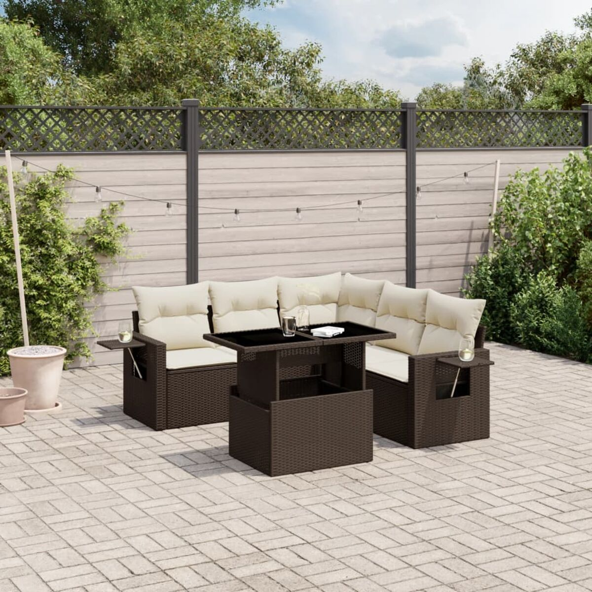 Dunkelbraunes Wicker-Ecksofa mit cremefarbenen Kissen und einem schwarzen Glastisch. Steinterrasse, Holzzaun und Pflanzen.