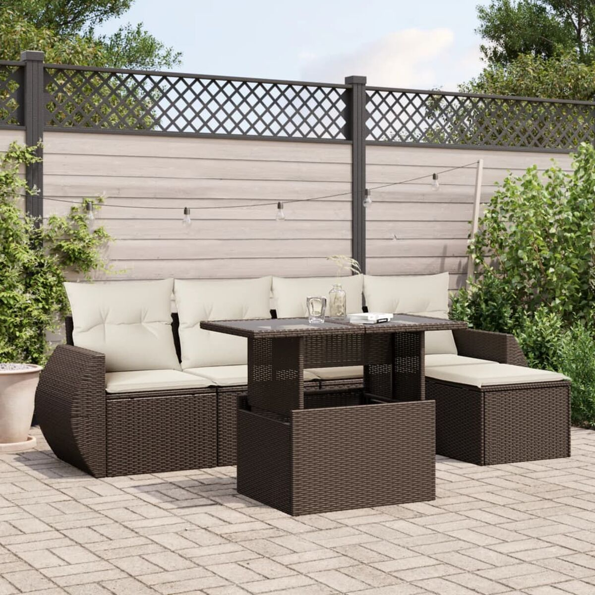 Set da esterno in vimini marrone scuro con cuscini color crema e tavolo. Patio in mattoni, recinzione in legno, verde.