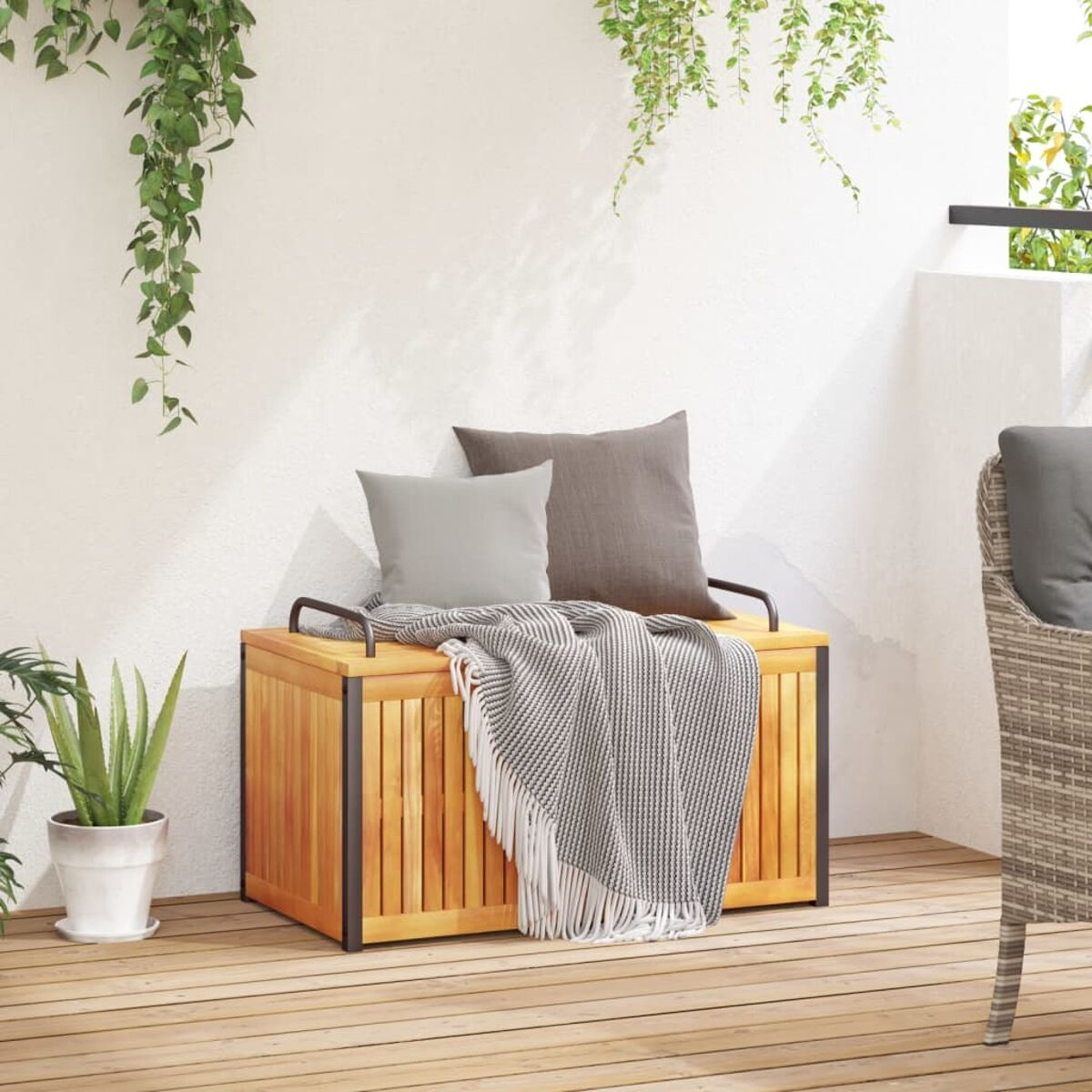 Holz-Außenbank mit Kissen und Decke. Pflanzen und ein teilweiser Korbstuhl sind sichtbar.
