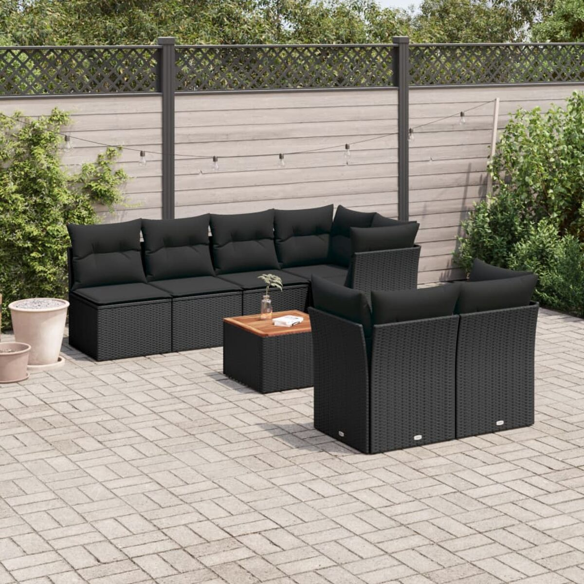 Schwarzes Rattan-Sofaset mit Kissen, Holztisch und Pflastersteinen. Grüne Pflanzen und Zaun im Hintergrund.