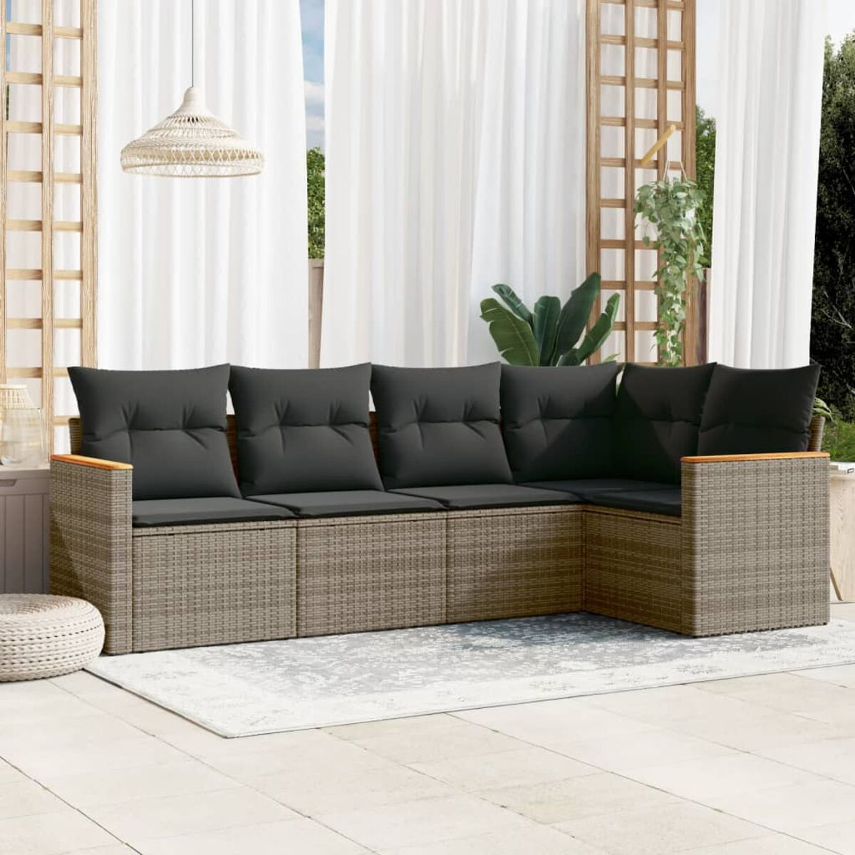 Graues Ecksofa aus Korbgeflecht mit schwarzen Kissen auf einer Terrasse. Ein weißer Teppich und Hocker.