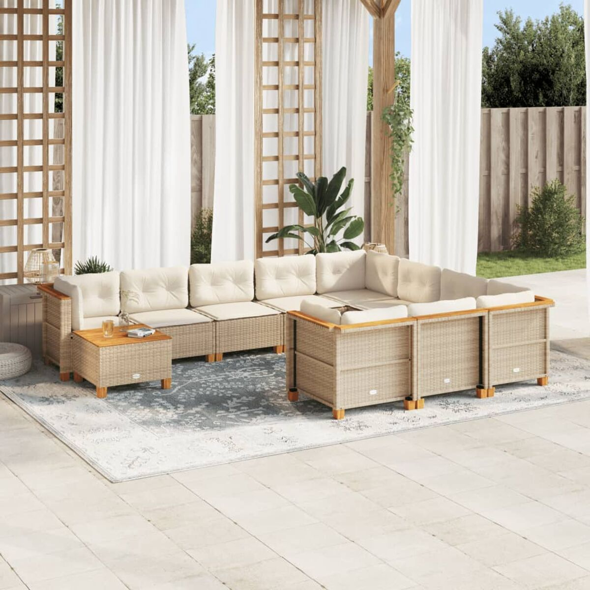 Set divani in vimini da esterno con cuscini beige, tavolino, su un tappeto fantasia. Tende bianche sullo sfondo.