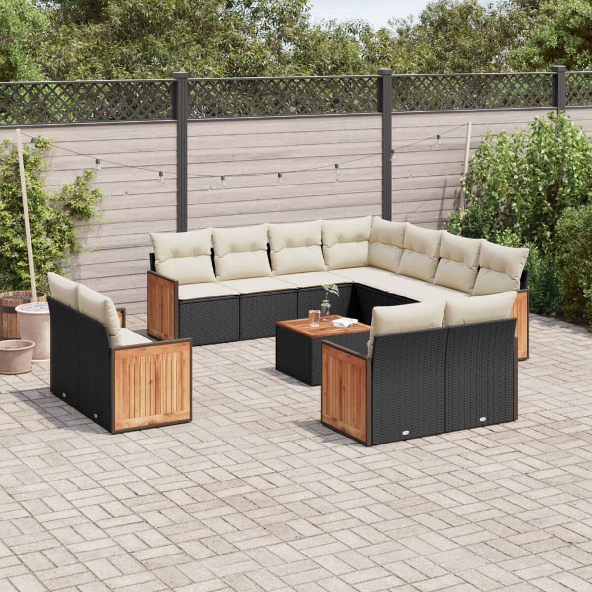 Schwarzes Rattan-Sofaset mit beigen Kissen und Holzakzenten. Auf einer gepflasterten Terrasse, Pflanzen, Zaun.