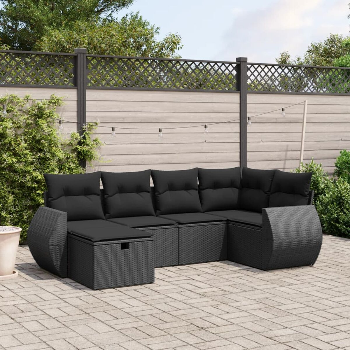 Schwarzes Outdoor-Ecksofa aus Korbgeflecht mit schwarzen Kissen, auf einer Backsteinterrasse, mit Zaun und Grün.