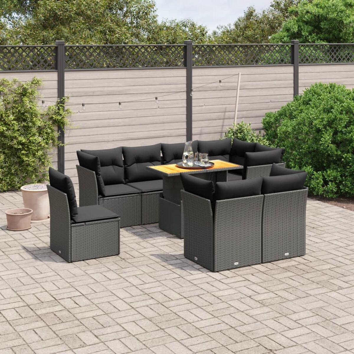 Graues Rattan-Gartenmöbel-Set mit schwarzen Kissen und Tisch. Ziegel-Patio, grüne Pflanzen.