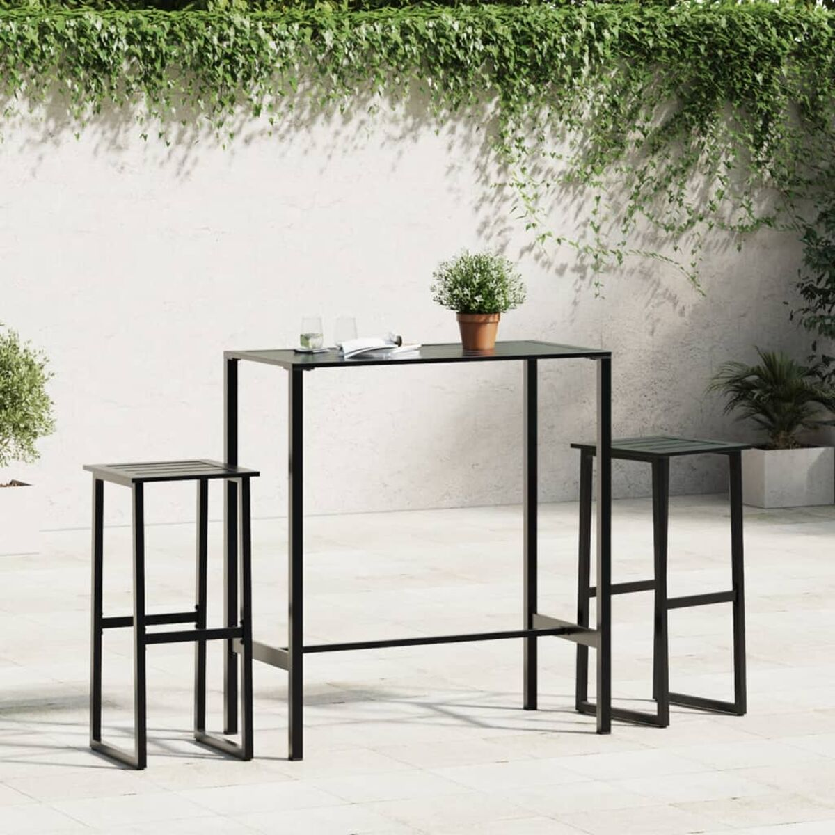 Un tavolo da bar in metallo nero con due sgabelli. Il tavolo e gli sgabelli sono su un patio bianco, con piante sullo sfondo.