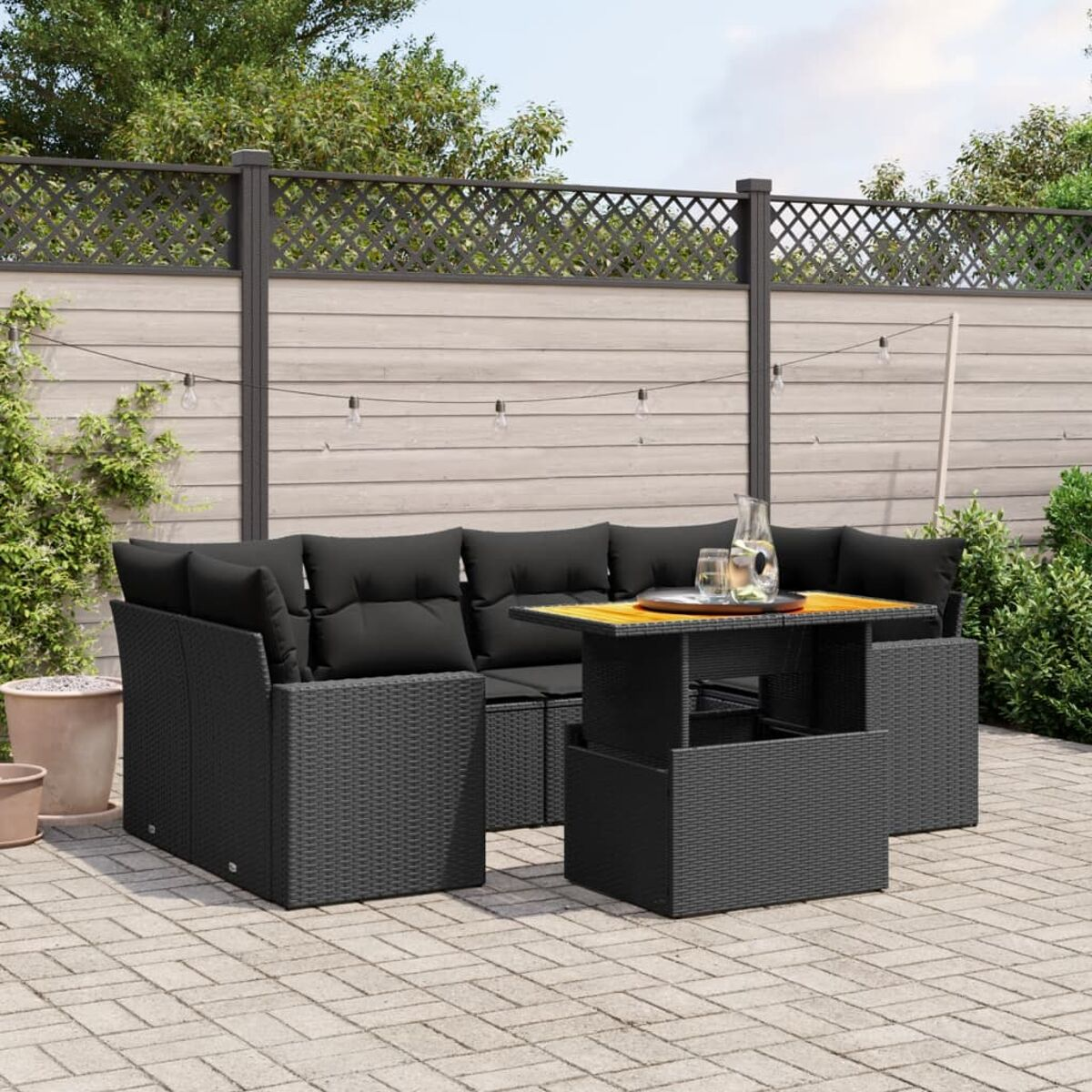 Schwarzes Rattan-Außensofa mit Kissen und einem Tisch. Lichter und Pflanzen im Hintergrund.