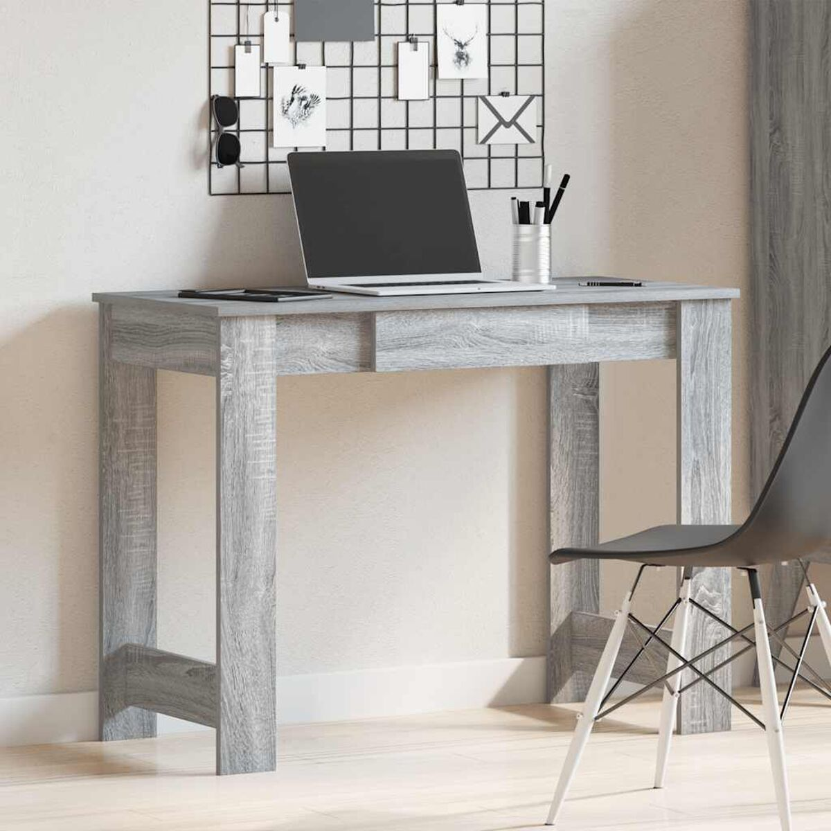 Ein grauer Schreibtisch mit Laptop, Stifthalter und Stuhl, mit einem Gitter an der Wand.