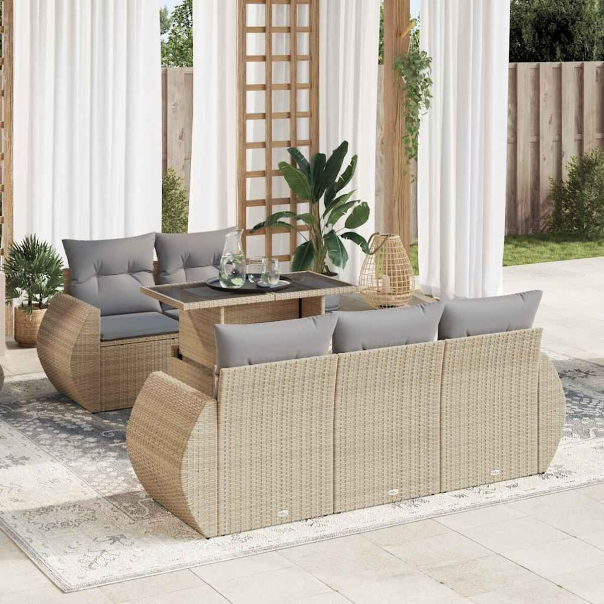 Set divano in rattan da esterno con tavolo, sedie e tende bianche in un giardino.