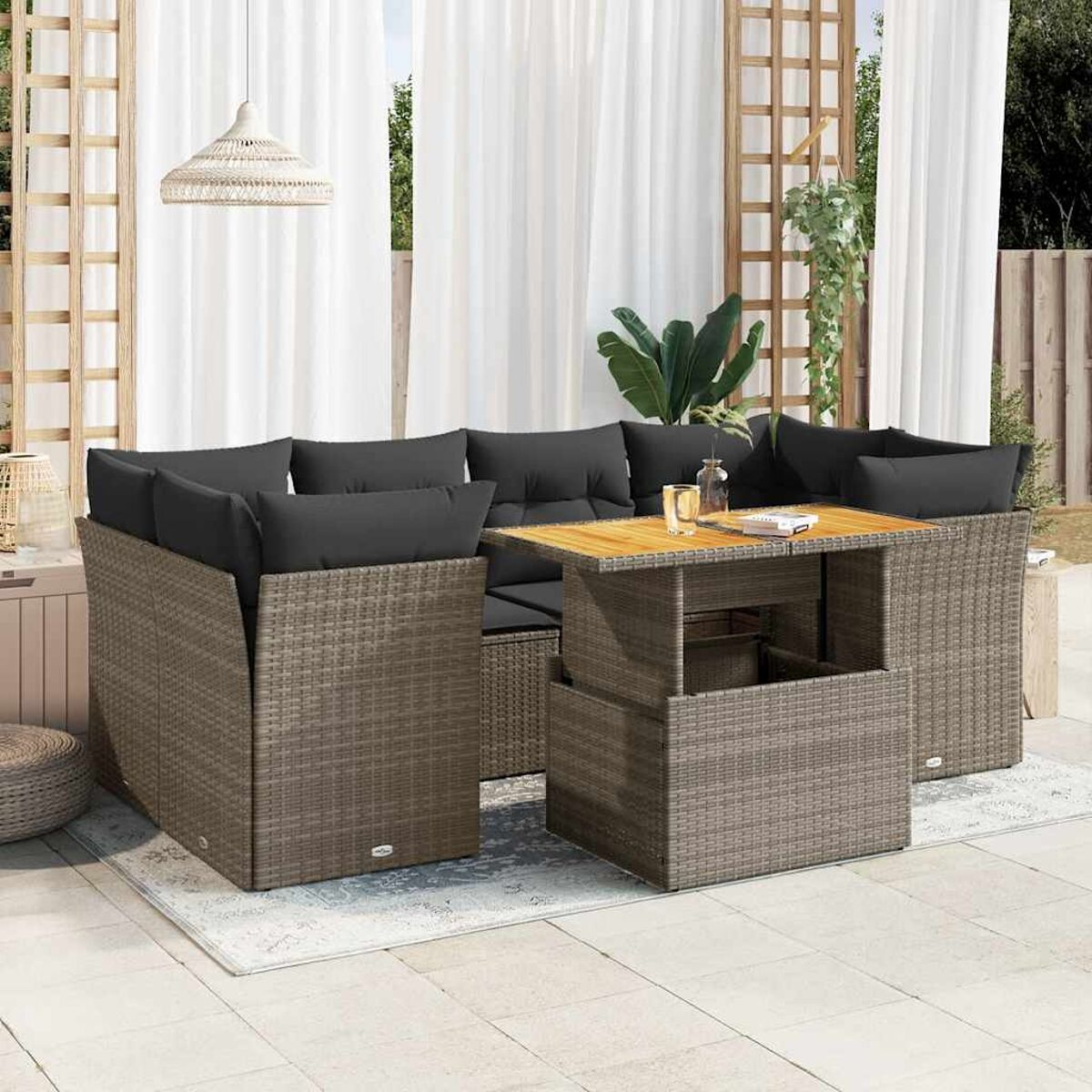 Set da patio in vimini con cuscini grigi. Include un tavolo e un divano angolare. Sfondo: tende bianche.