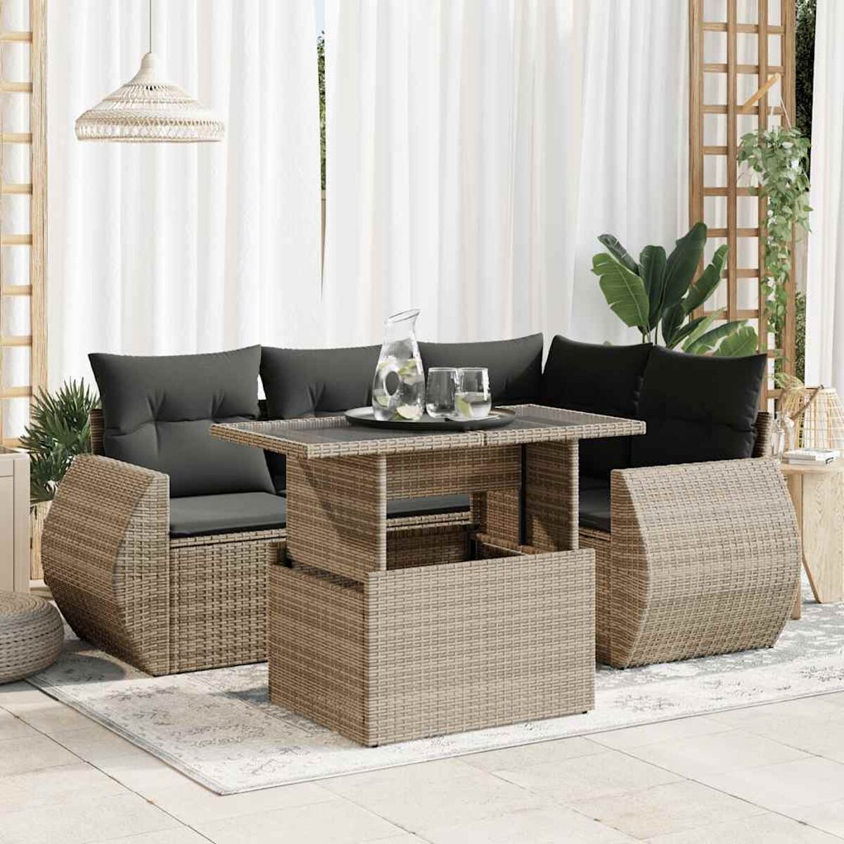 Set di mobili in rattan su un tappeto: divano, tavolo e sedie con cuscini, con tende e piante sullo sfondo.