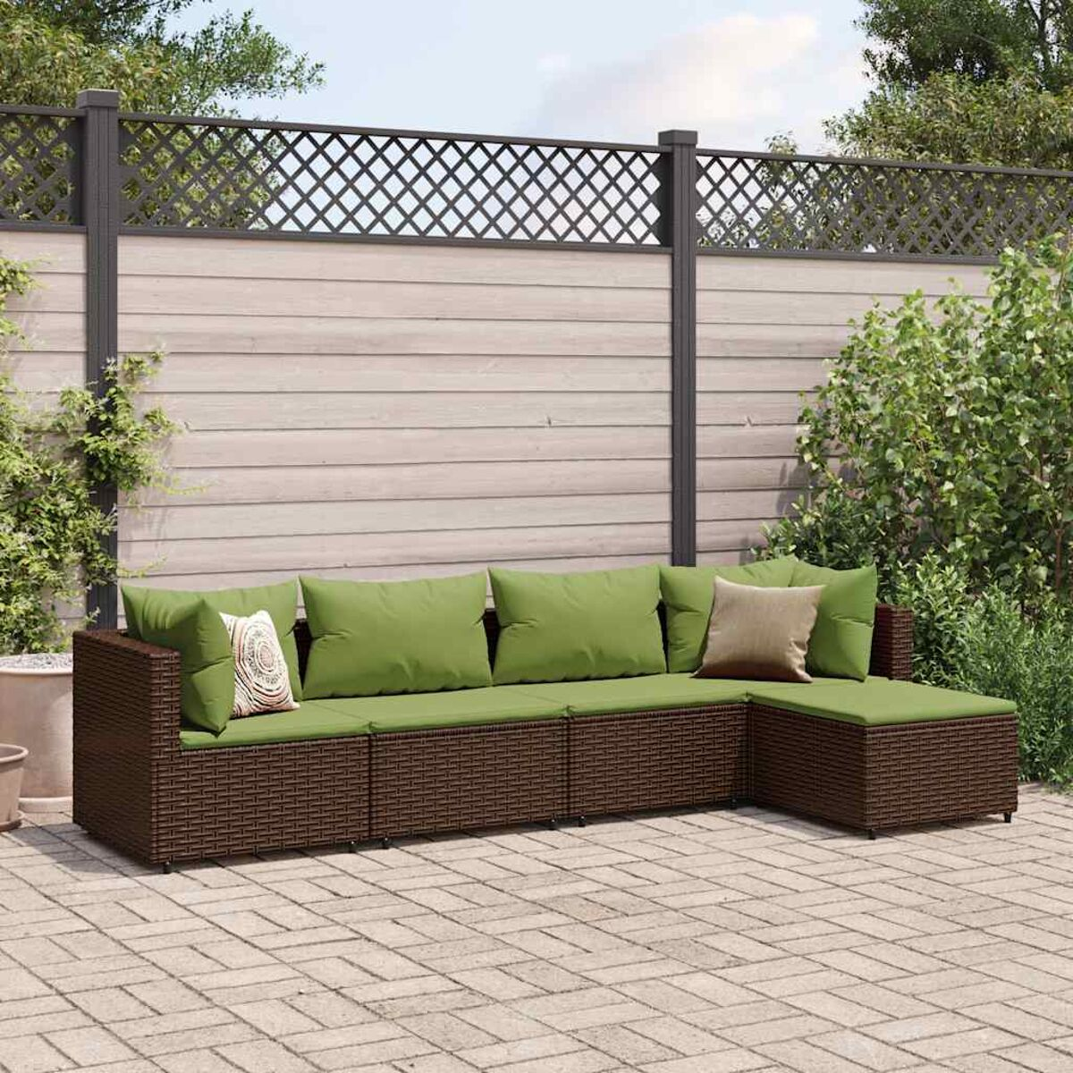 Divano componibile in vimini marrone con cuscini verdi. Su patio in mattoni con recinzione in legno e cespugli verdi.