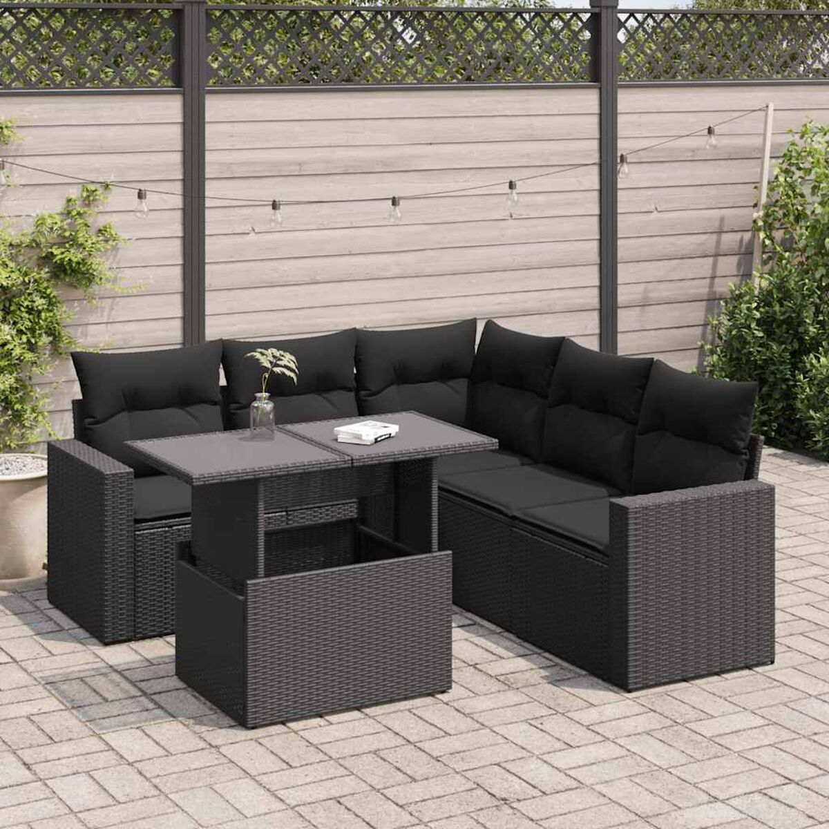 Divano angolare in rattan nero con cuscini neri, un tavolo grigio. Ambiente patio con verde e recinzione.