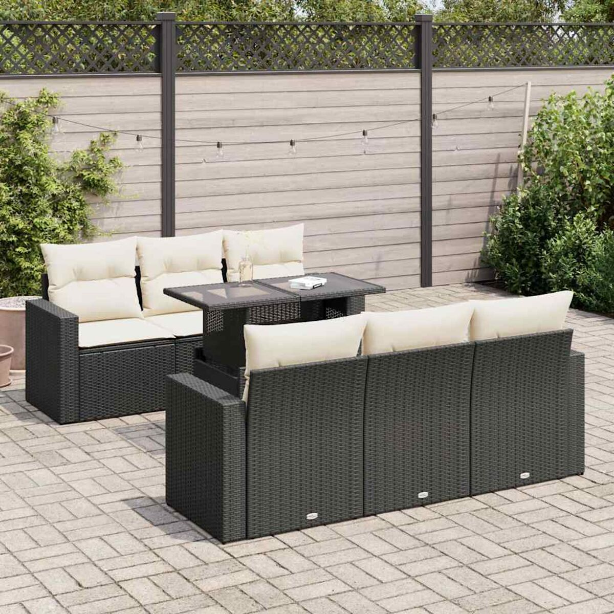 Set di divani da esterno in vimini nero con tavolo e cuscini su un patio. Recinzione e piante sullo sfondo.