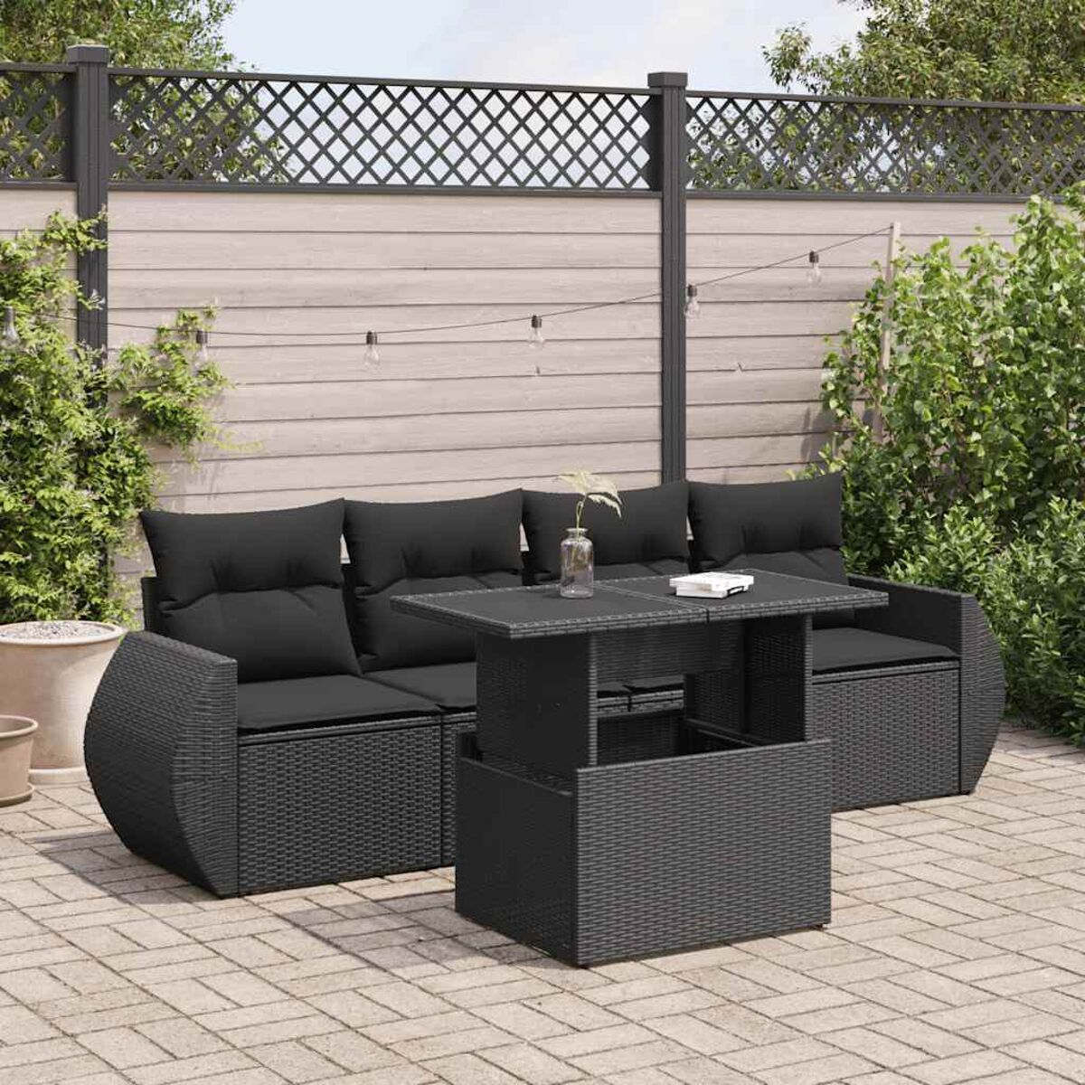 Set patio nero con divano, tavolo e sedie. Cuscini scuri. Recinzione in legno e piante sullo sfondo.