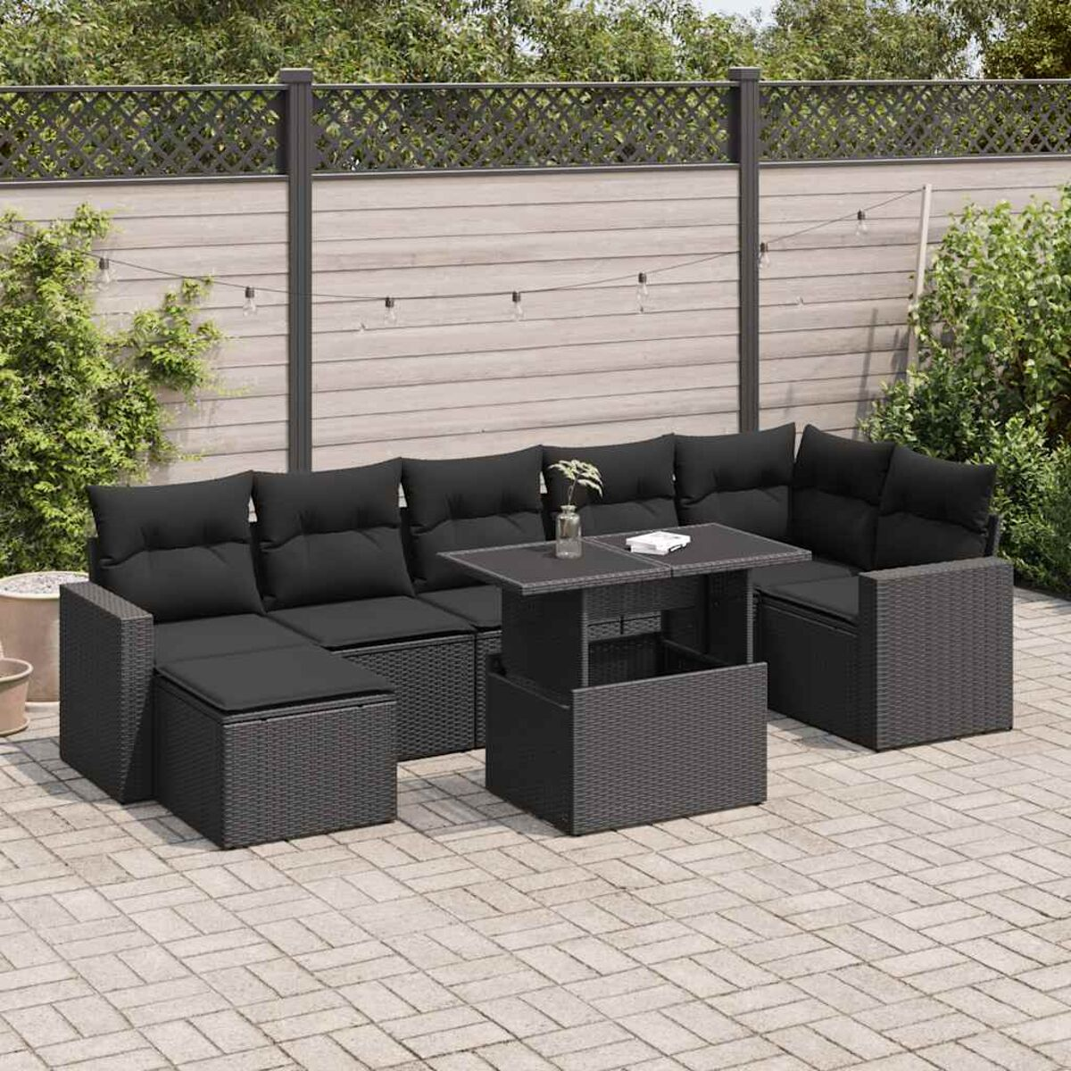 Set da patio con cuscini neri e tavolo. Include piante e una recinzione.