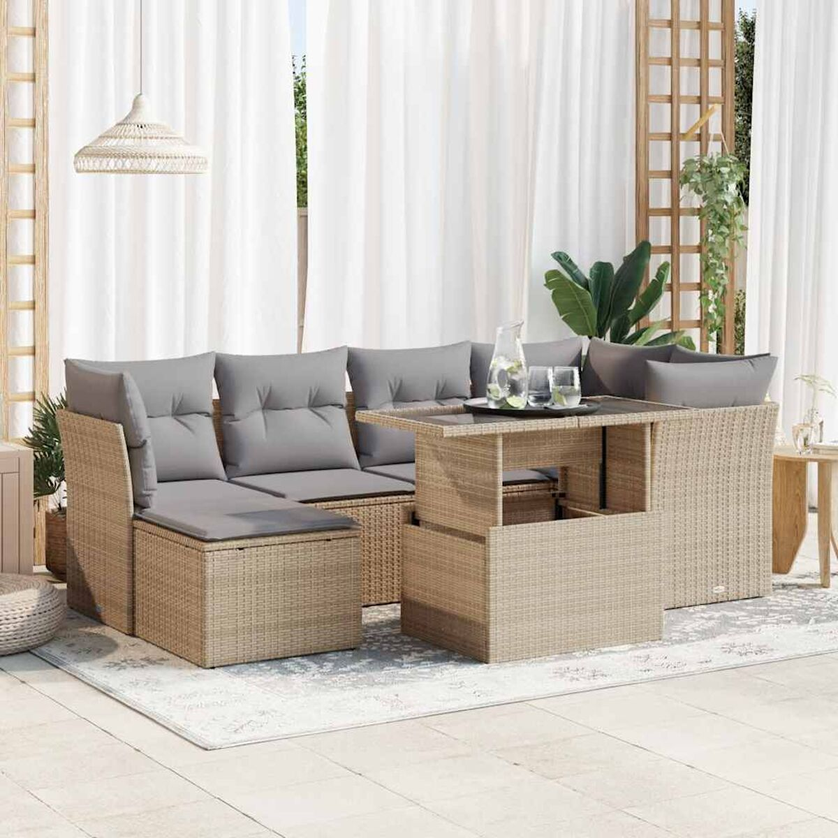 Set divano componibile da esterno in rattan beige con cuscini grigi e un tavolo. Tende bianche, tappeto e fogliame sullo sfondo.