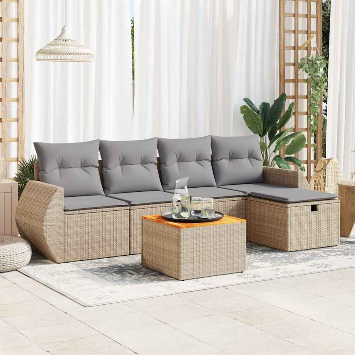 Divano in rattan con cuscini grigi e tavolo in legno con bicchieri e caraffa. Tende bianche sullo sfondo.