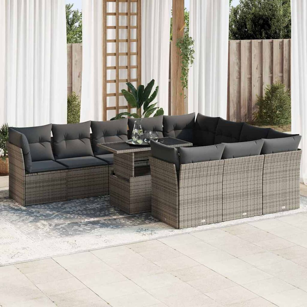 Set divano da esterno in vimini grigio con cuscini grigi, tavolo. Su un patio, con tende e piante sullo sfondo.