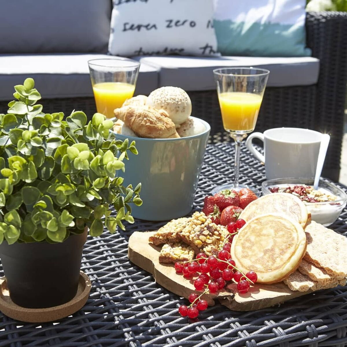 Frühstück auf einem Tisch: Orangensaft, Gebäck, Obst und Müsli. Umgebung im Freien mit Kissen.