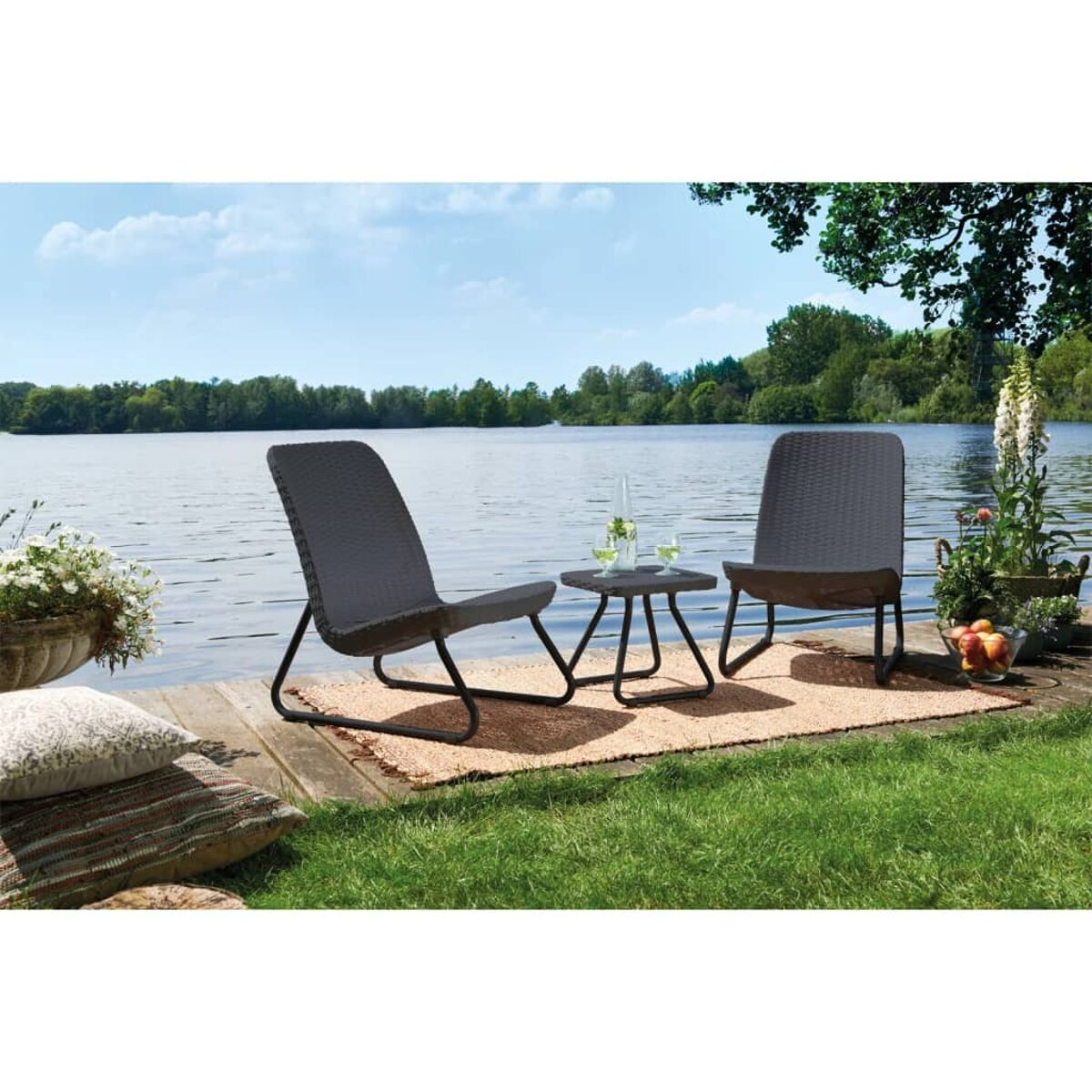 Szene am Seeufer: zwei Stühle, kleiner Tisch, Teppich auf Holzsteg, mit Wasser, Bäumen und Himmel im Hintergrund.