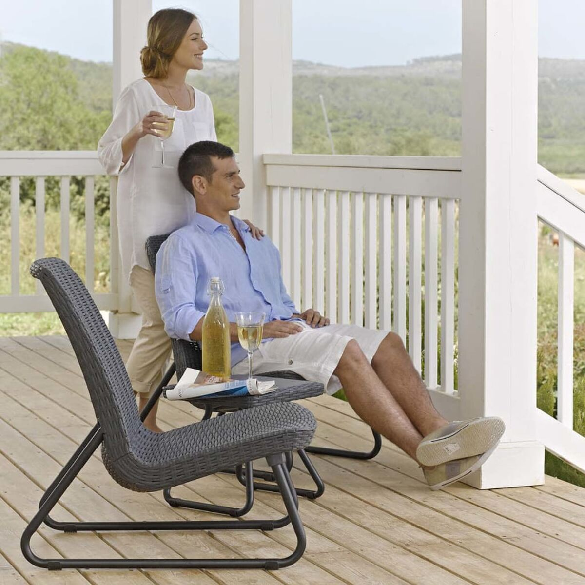 Ein Mann und eine Frau genießen Getränke auf einer Veranda. Ein Tisch mit Flasche und Buch ist in der Nähe.