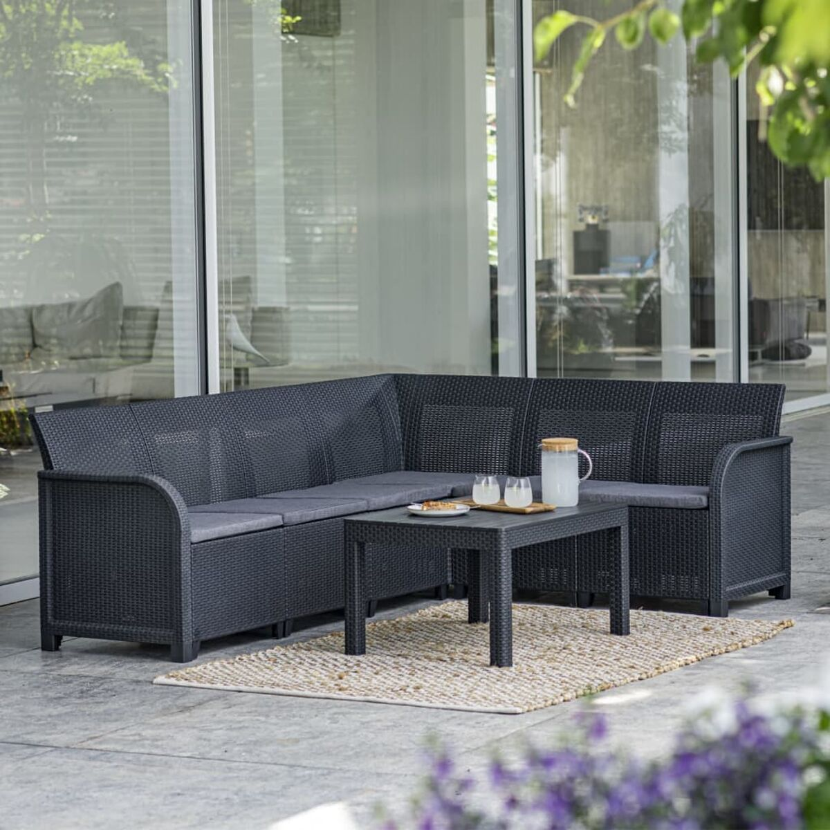 Schwarzes Rattan-Outdoor-Sitzgruppe mit Tisch und Teppich. Haus und Pflanzen im Hintergrund.