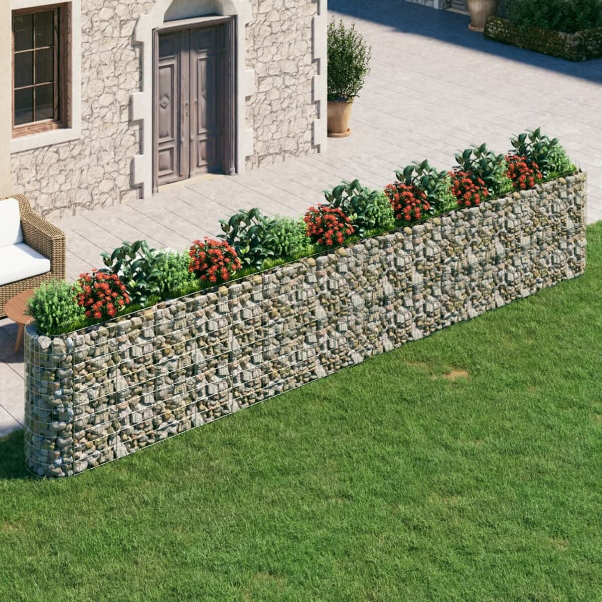 Steinmauer mit Pflanzen und roten Blumen. Grünes Gras im Vordergrund, Haus im Hintergrund.