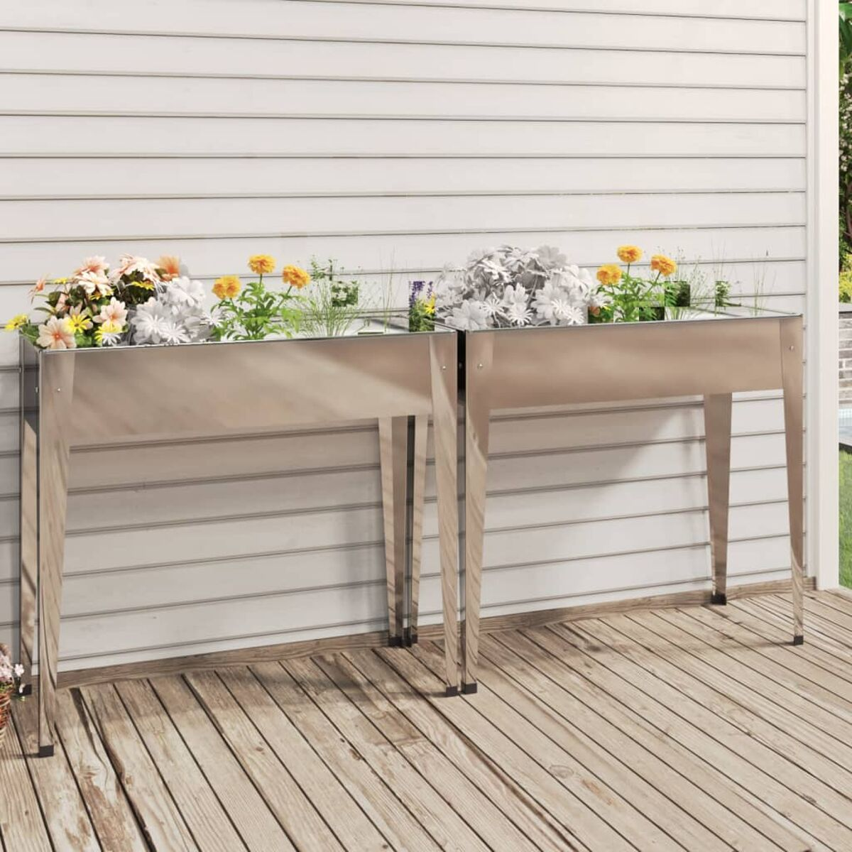 Zwei silberne Metallpflanzgefäße mit Blumen auf einer Holzterrasse vor einer weißen Wand.