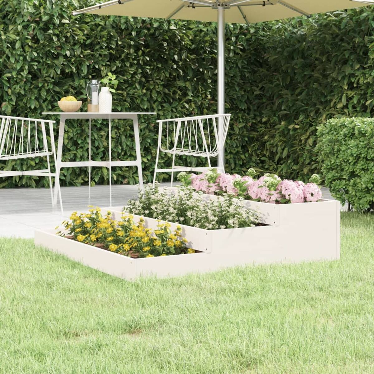 Weißer Stufenpflanzer mit Blumen und Grün. Im Hintergrund ein Tisch und ein Sonnenschirm.