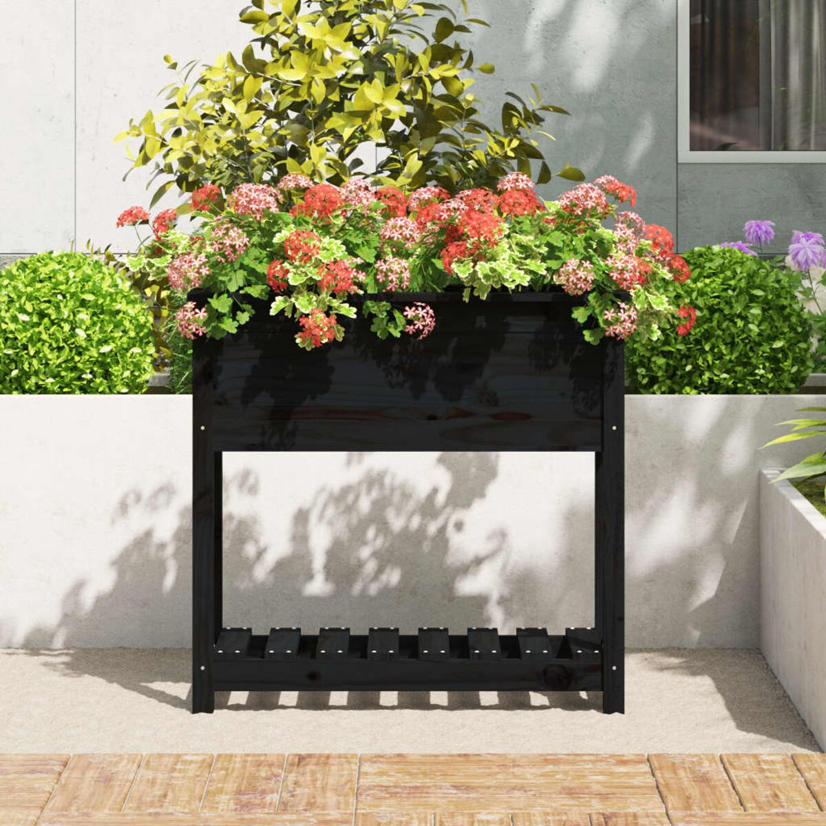 Schwarzer Holzpflanzkasten mit roten Blumen, auf einer hellen Terrasse, mit Gebüsch im Hintergrund.