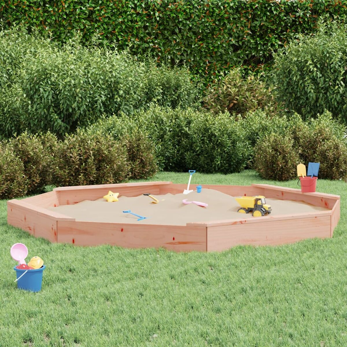Sechseckige Holz-Sandkiste mit Sand und Spielzeug gefüllt. Grünes Gras umgibt die Sandkiste.